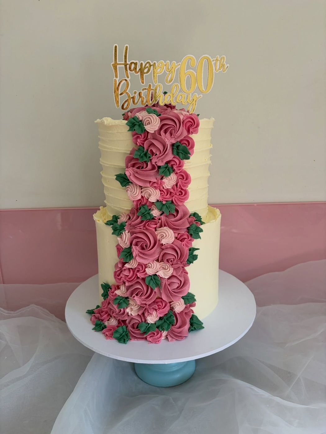 A two-tier cream cake with a pink buttercream rose cascade and a gold Happy 60th Birthday topper by Lakey's Bakes