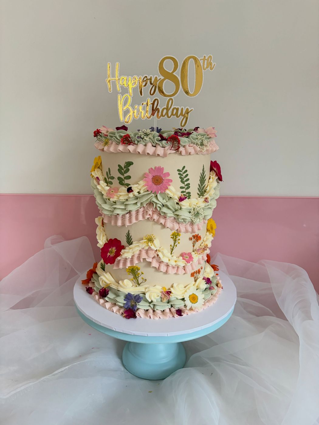 A two-tier floral cake with a gold Happy 80th Birthday topper